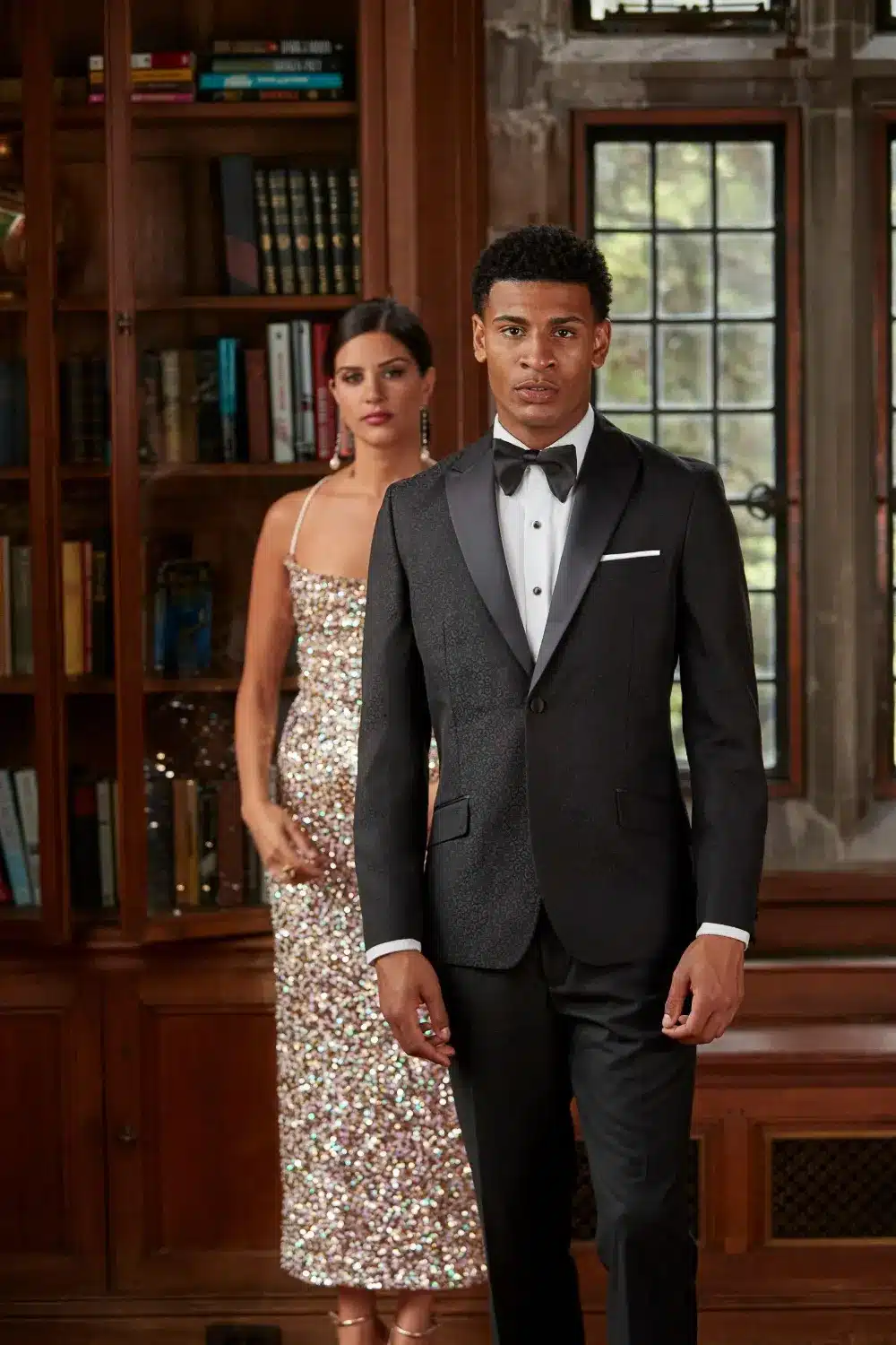 Special Occasions - Black Tie Tuxedo Man in a black tuxedo with bow tie and his date in a sequin gown in a wood-paneled mansion library - special occasion tuxedo rental at Minsky Formal Wear, Richardson